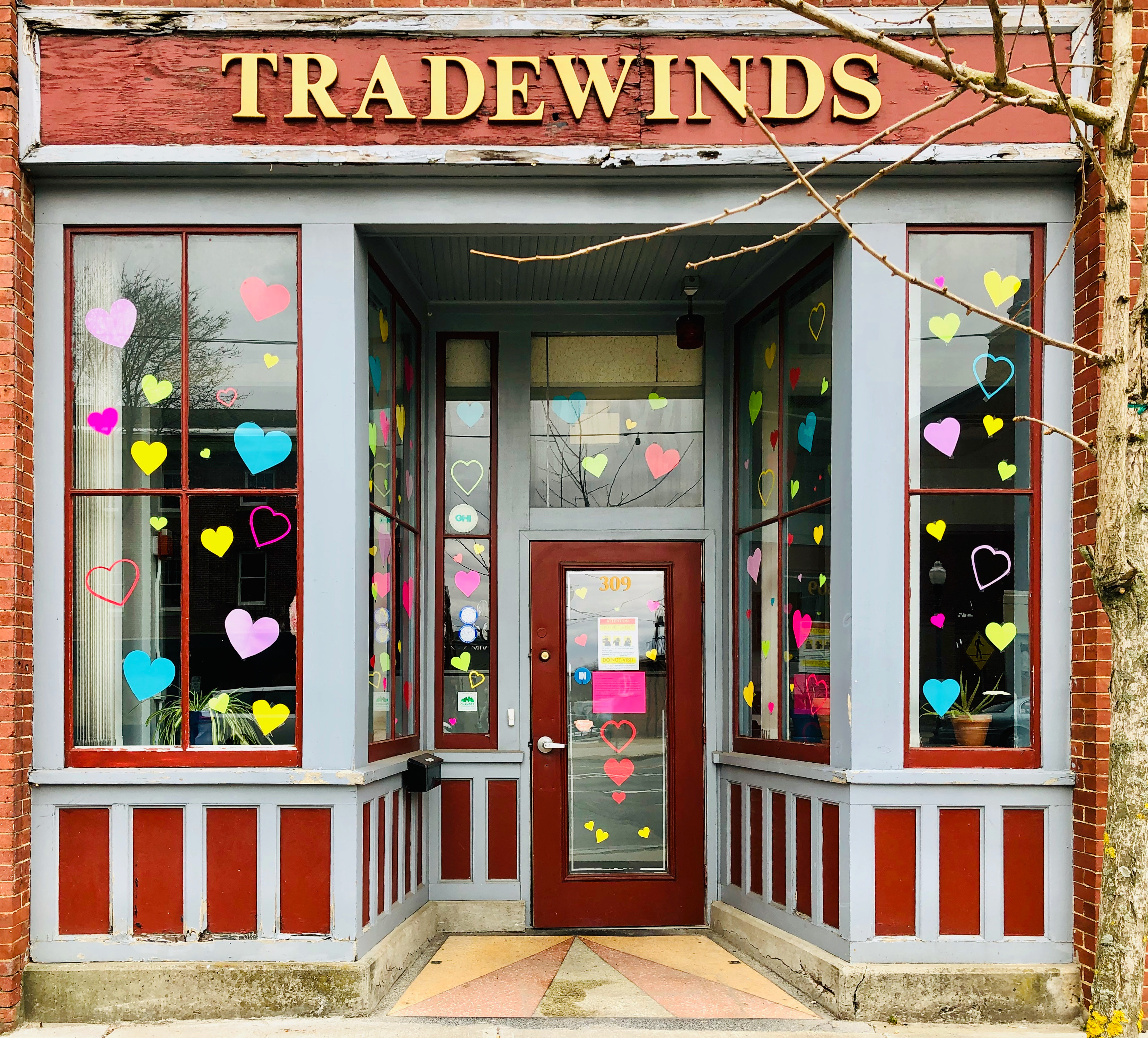 Image of the a red brick building with golden letters on top saying "Tradewinds" in all capital letters. The window panes are decorated with pink, blue, and yellow hearts.