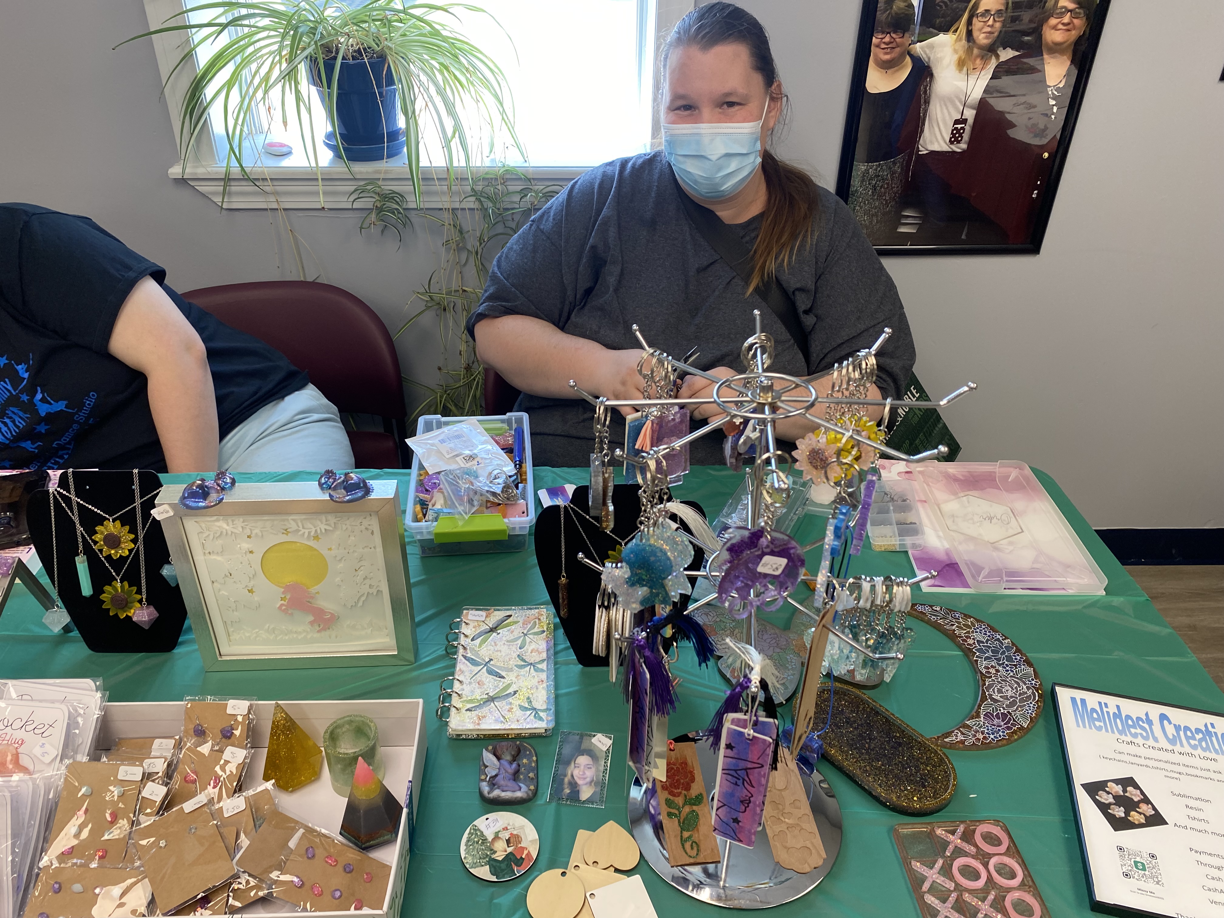 Image of a white female with a gray sweater and brown hair in a ponytail. She is wearing a blue mask and is sitting in front of a green table. The table has an assortment of jewelry, decorations, and other crafts. Another person is on the left side leaning out of view.